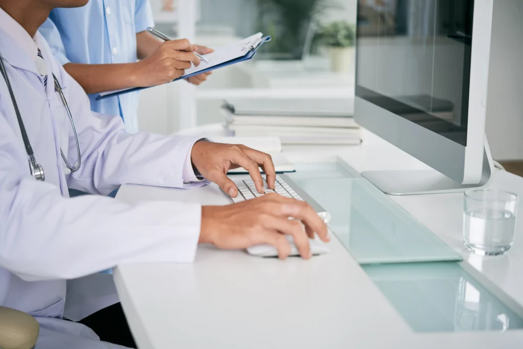 Médico escribiendo en computadora con estetoscopio, asistente con clip.