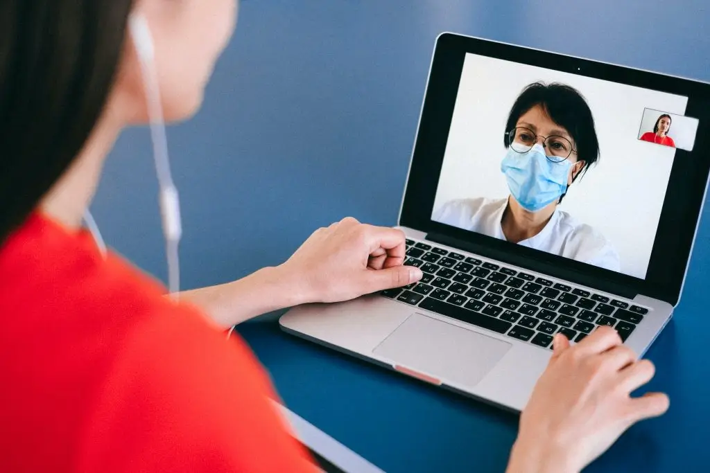 Mujer en videoconferencia con otra mujer con mascarilla en pantalla de laptop.