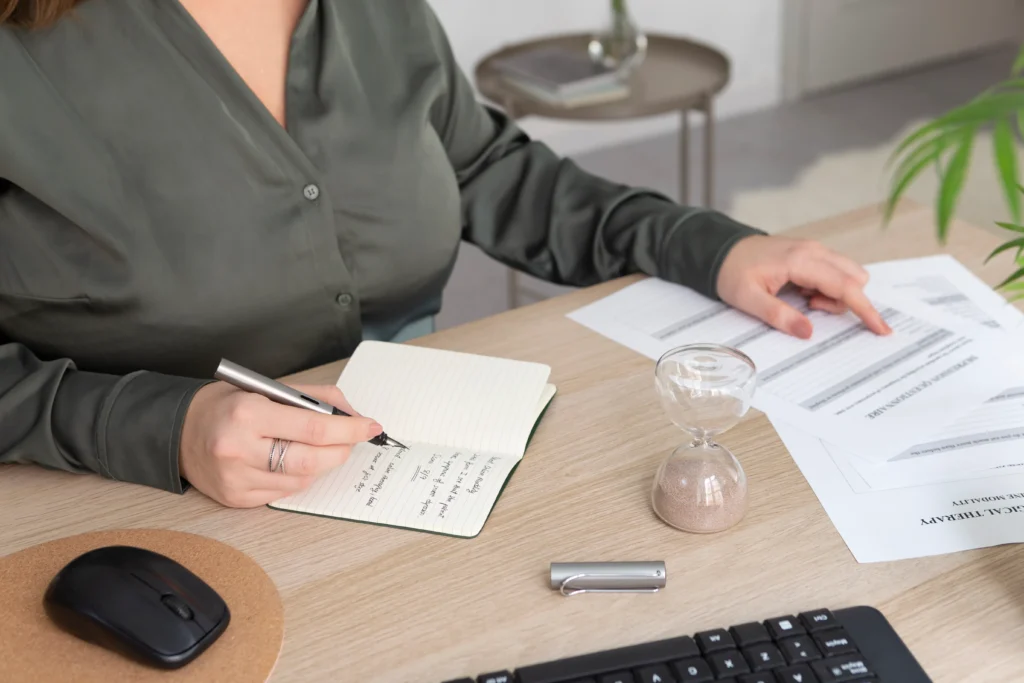 Mujer escribiendo en un cuaderno con documentos y temporizador en la mesa de trabajo.