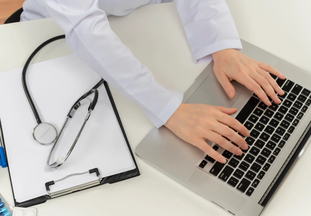 Médico trabajando en laptop con estetoscopio sobre documentos.